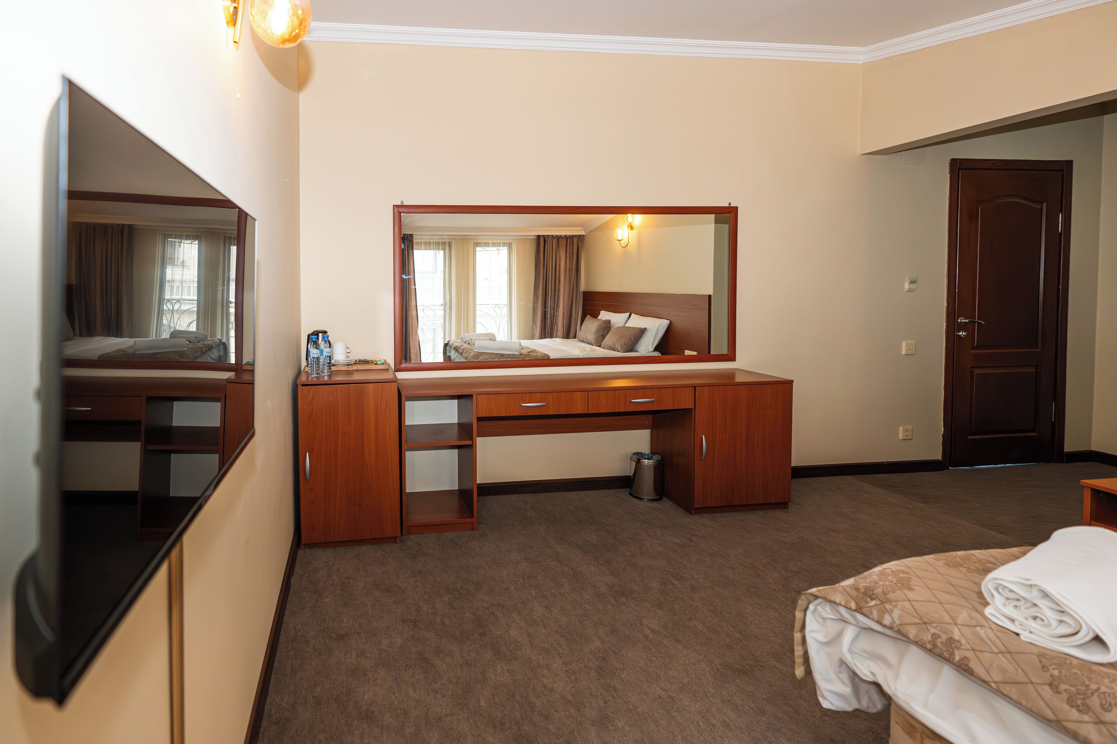 A hotel room with a bed, a desk, a mirror, a trash can, and a door.