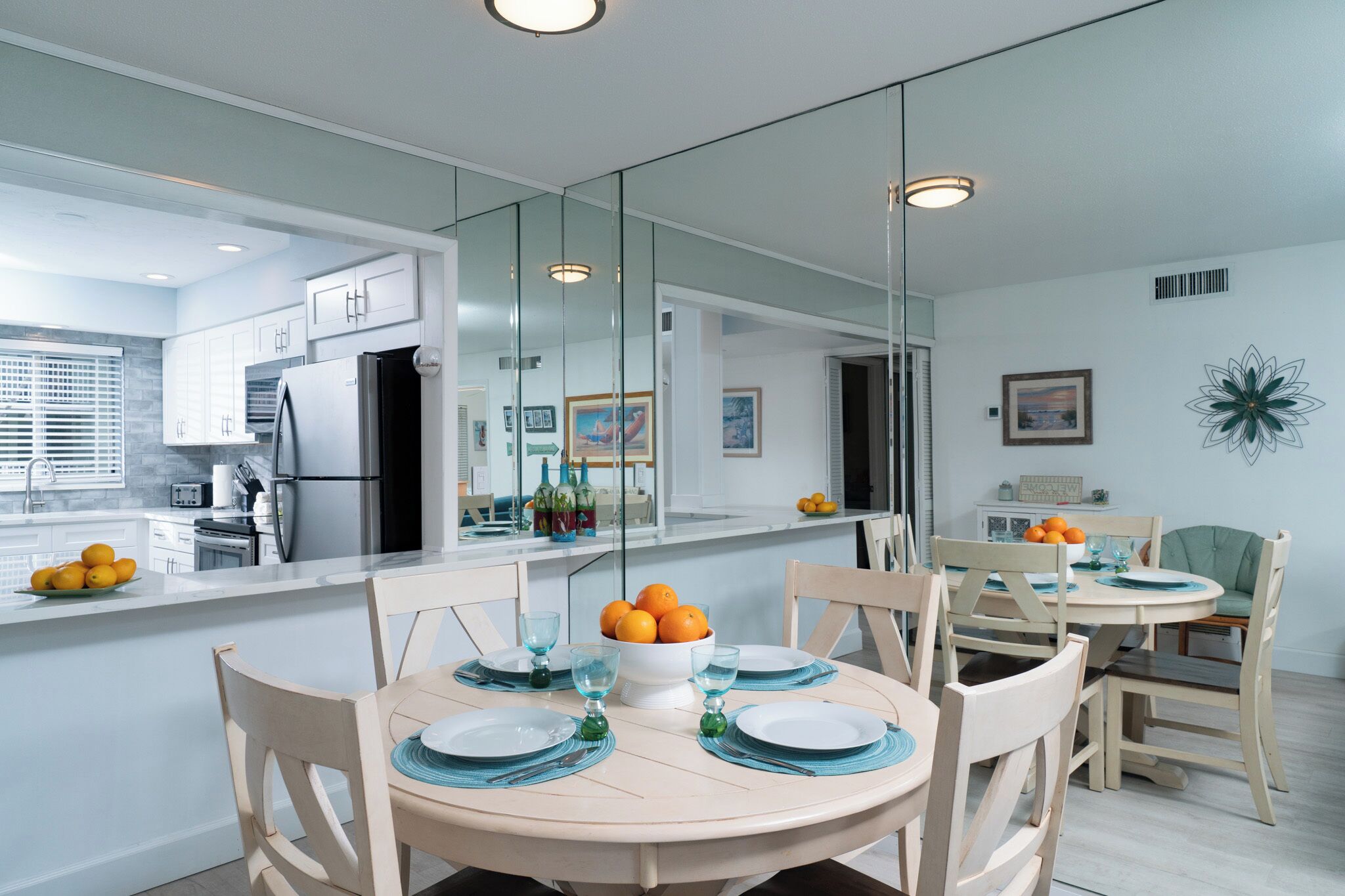 A modern kitchen with a large island, a dining area with a round table and chairs, and a mirrored wall reflecting the kitchen.