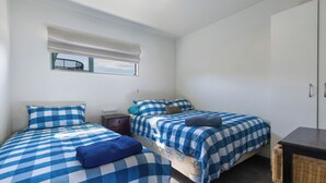 A room with two beds, each with a checkered blue and white bedspread, a small table, a wicker chair, and a window with a view.