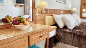A hotel room with a wooden desk, a bowl of fruit, a bed with a brown headboard, and a nightstand with a lamp.