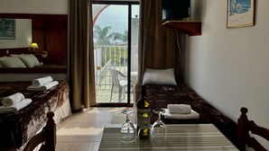 A hotel room with a bed, a small table with a bottle and glasses, a TV on the wall, and a balcony with a view.