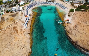 Une vue aérienne d’une plage avec une étendue d’eau d’un bleu limpide, une rangée de parasols et un quai où sont amarrés des bateaux.