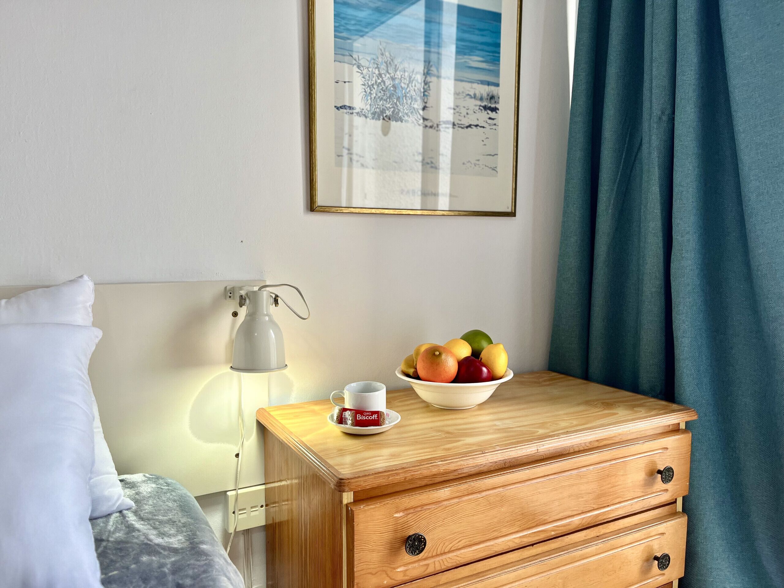 A neatly arranged hotel room with a bed, a bedside table with a lamp, a bowl of fruit, and a framed picture on the wall.
