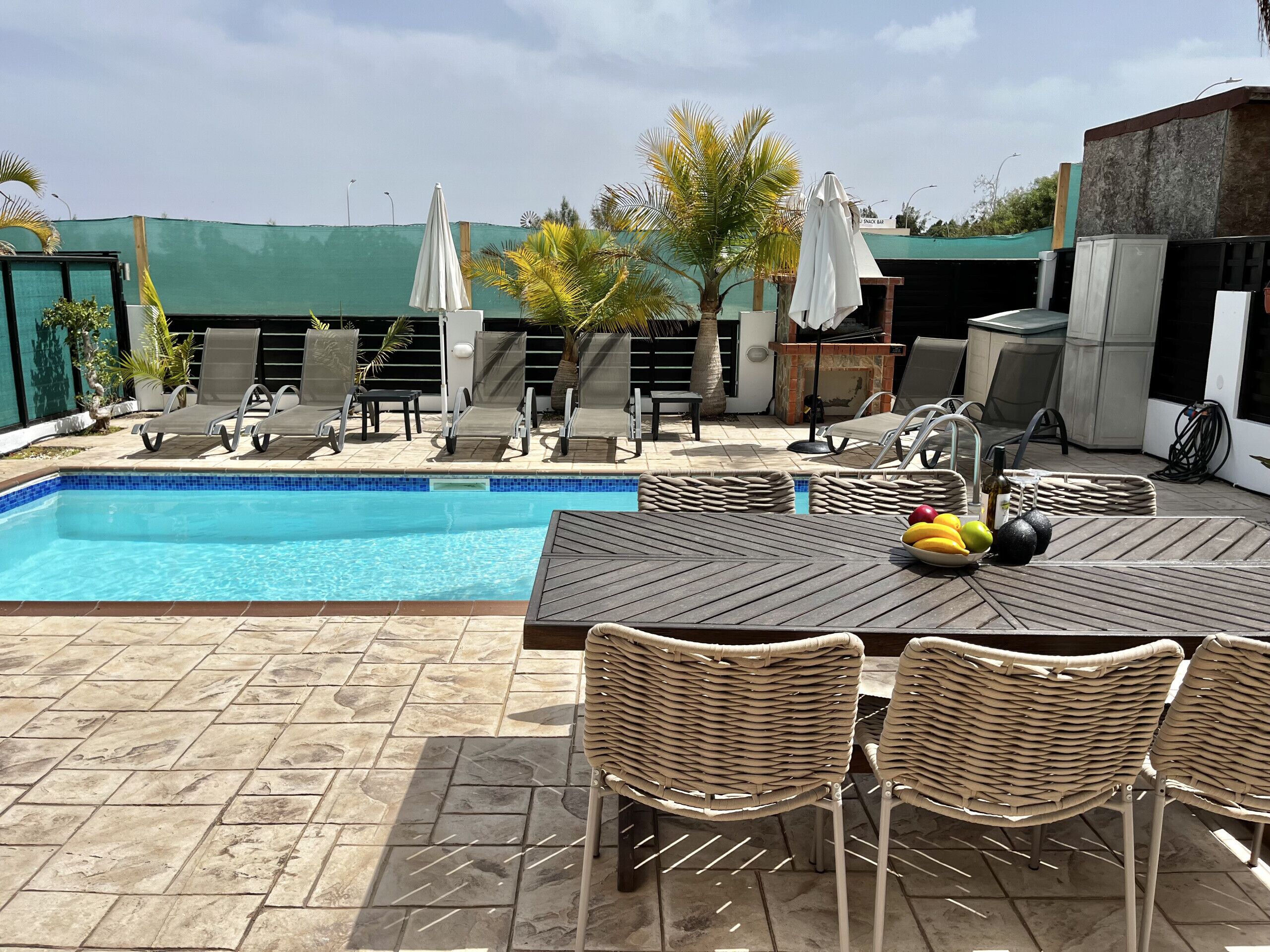 Zona junto a la piscina con piscina, sillas de descanso, una mesa con frutas y sombrillas.