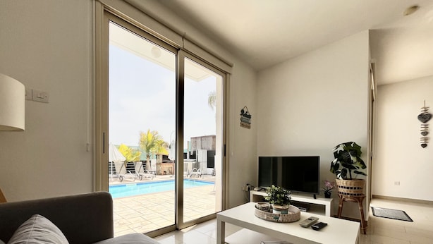 A modern living room with a grey sofa, a flat-screen TV, a small table with a plant, and a sliding glass door leading to an outdoor pool area.