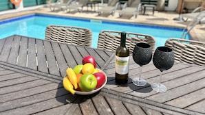 A poolside table with a bowl of fruit, a bottle of wine, and two glasses.