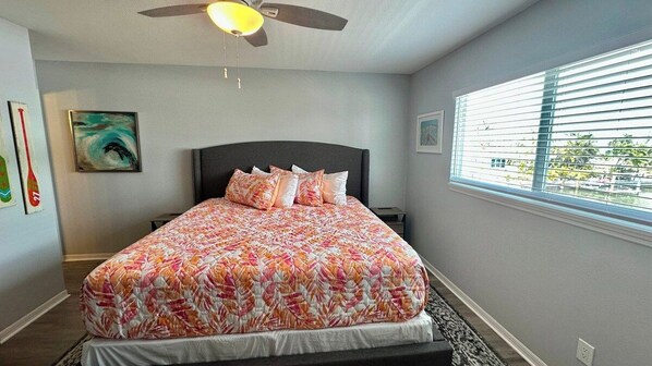 A bedroom with a bed, a ceiling fan, a window with blinds, and artwork on the walls. - The Lighthouse (Fort Myers Beach)