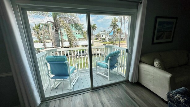 A room with a sliding glass door leading to a deck with two blue chairs and a white fence. - The Lighthouse - Steps from Beach (Fort Myers Beach)