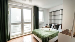 A modern bedroom with a large window, a bed with a green bedspread, a bedside table with a tray, and a built-in bookshelf. - Poznań Grunwald - 51m2, Balcony - by Rentujemy (Poznan)