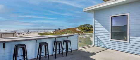A balcony with three black bar stools, a light blue building, and a view of the beach and ocean.