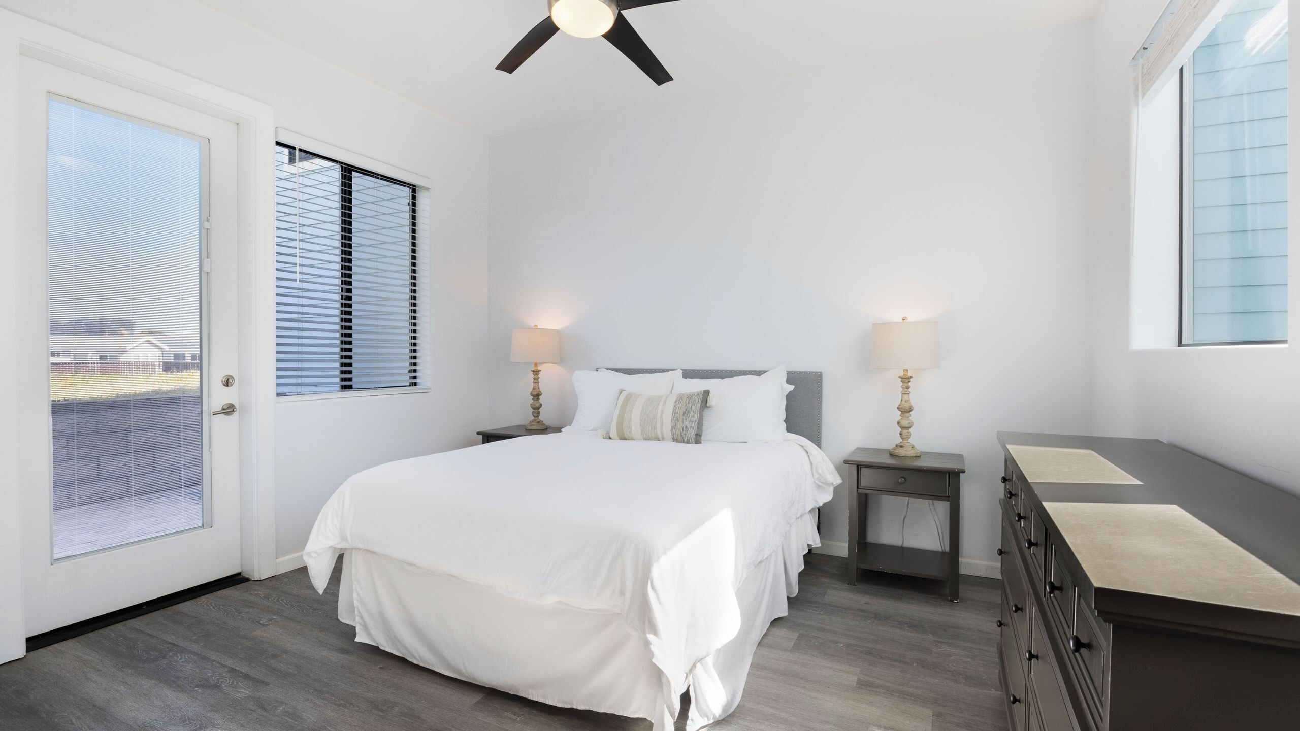 Un dormitorio con una cama, dos mesitas de noche con lámparas, un aparador, un ventilador de techo y una puerta con persianas.