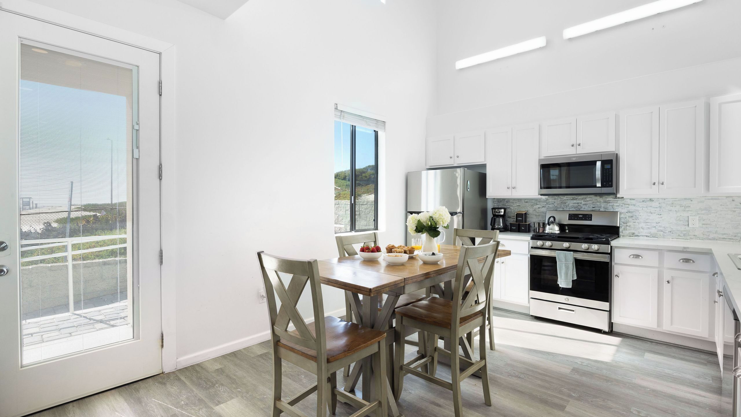 A modern kitchen with white cabinets, a stainless steel refrigerator, a microwave, and a stove. There is a dining table with chairs and a vase of flowers.