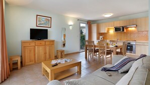 Een woonkamer met een bank, salontafel, tv en houten kasten. Er is een eethoek met een tafel en stoelen, en een keuken met een magnetron en oven.