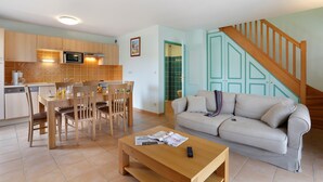 Een moderne keuken met houten kastjes, een eettafel en stoelen. Er is een zithoek met een bank en salontafel. Een trap met houten leuningen leidt naar een hoger niveau.