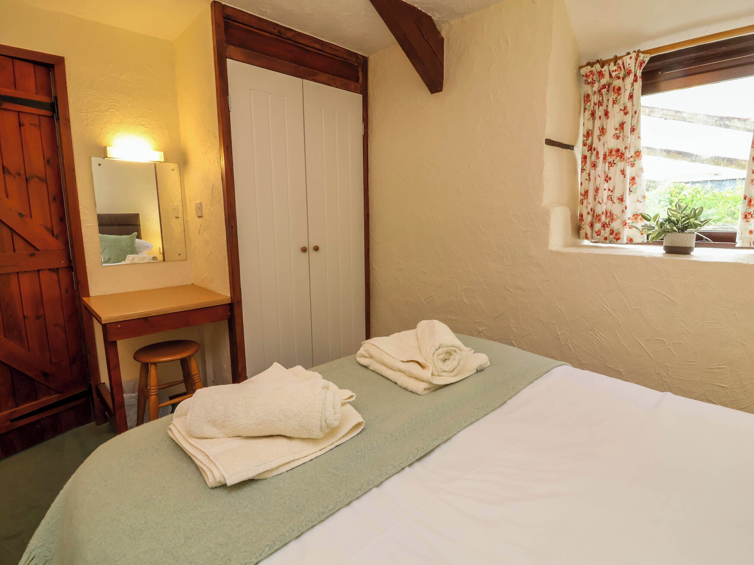 Ein Schlafzimmer mit einem Bett, einem Holztisch, einem Stuhl, einem Spiegel, einem Kleiderschrank und einem Fenster mit Vorhängen.