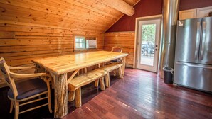 A wooden interior with a dining table and benches, a refrigerator, and a door leading outside.