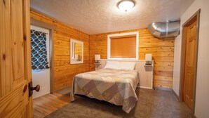 A cozy bedroom with wooden walls, a bed with a patterned bedspread, a window with blinds, and a ceiling vent.