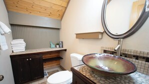 A bathroom with a wooden ceiling, a white toilet, a dark wooden cabinet, a glass sink, and a tiled backsplash.
