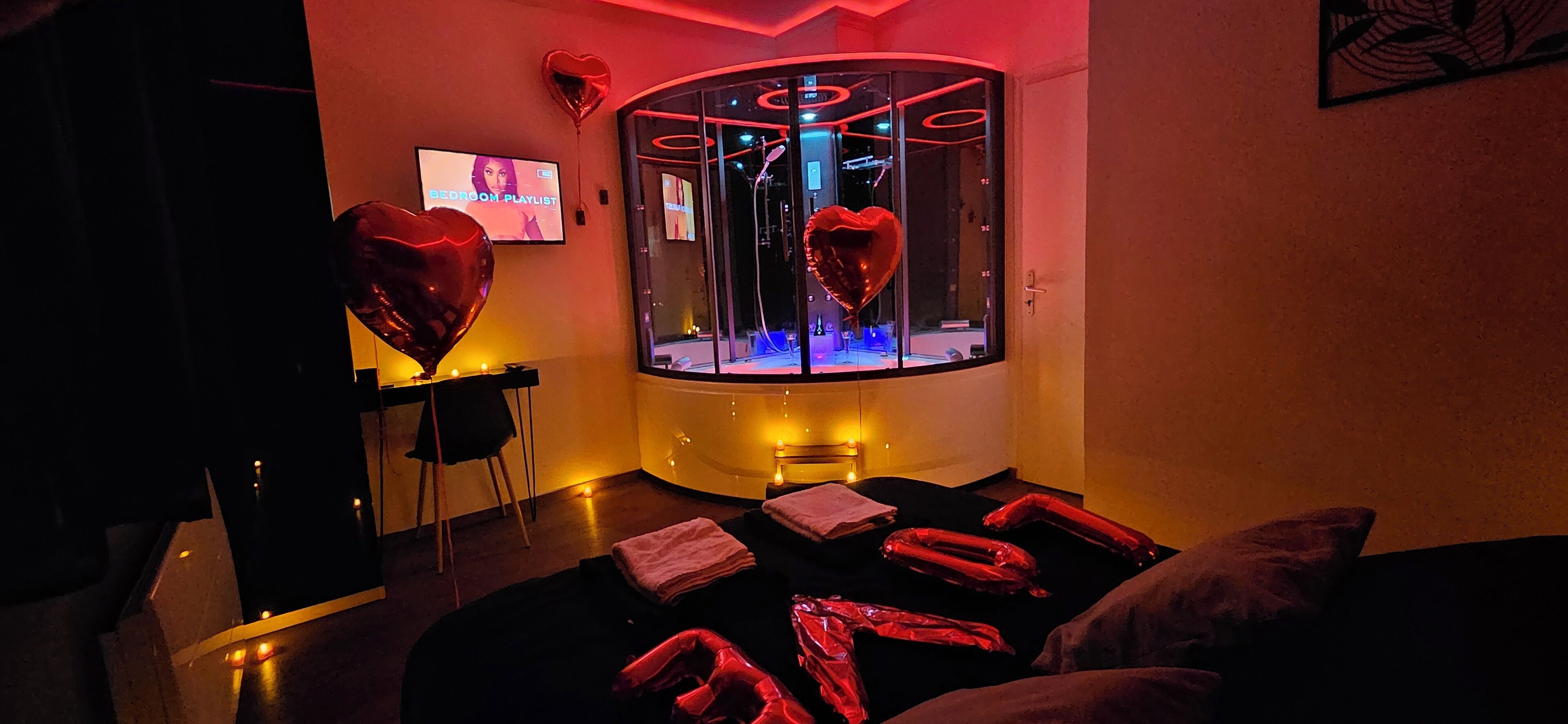 Ett svagt upplyst rum med en massagebänk, hjärtformade ballonger och en tv-skärm som visar en spellista med romantisk musik.