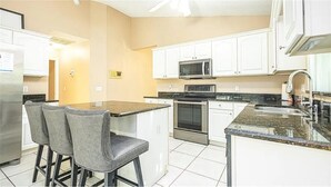 A modern kitchen with white cabinets, a microwave, an oven, and a granite countertop. There are two bar stools and a refrigerator. - Luxe Lakeside Escape (Davenport)