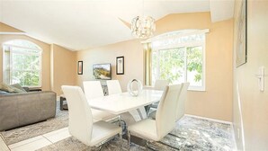 A modern dining area with a white table and chairs, a chandelier, and large windows with views of greenery. - Luxe Lakeside Escape (Davenport)