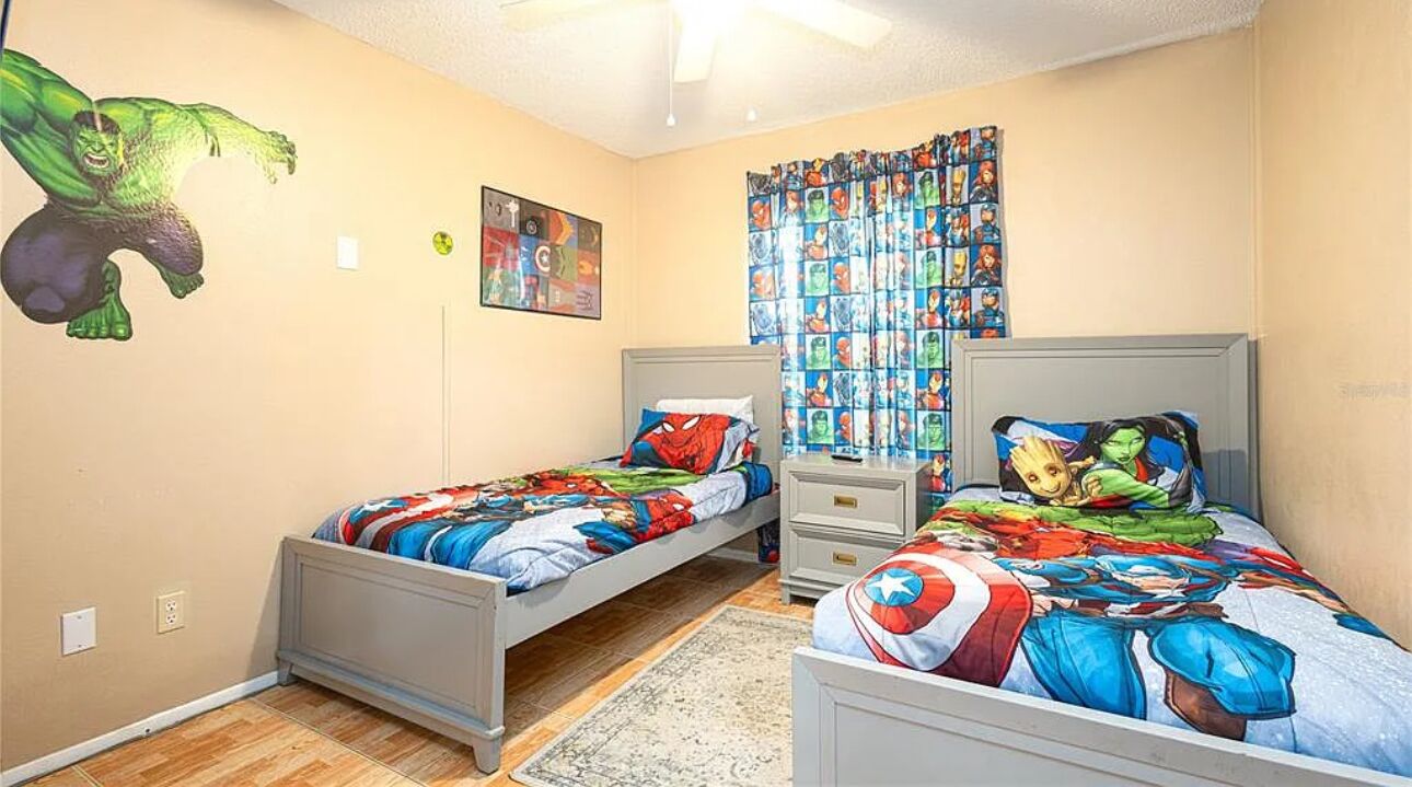 A bedroom with two single beds, a ceiling fan, a window with curtains, and a wall with a superhero mural.