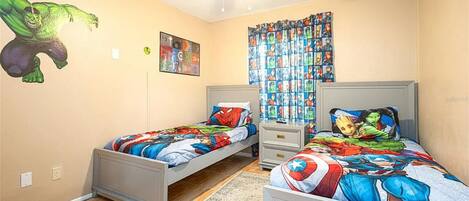A bedroom with two single beds, a ceiling fan, a window with curtains, and a wall with a superhero mural.
