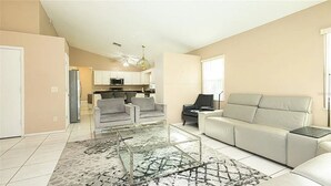 A spacious living room with a glass coffee table, two armchairs, and a sofa. A kitchen is visible in the background with white cabinets and a microwave. - Luxe Lakeside Escape (Davenport)