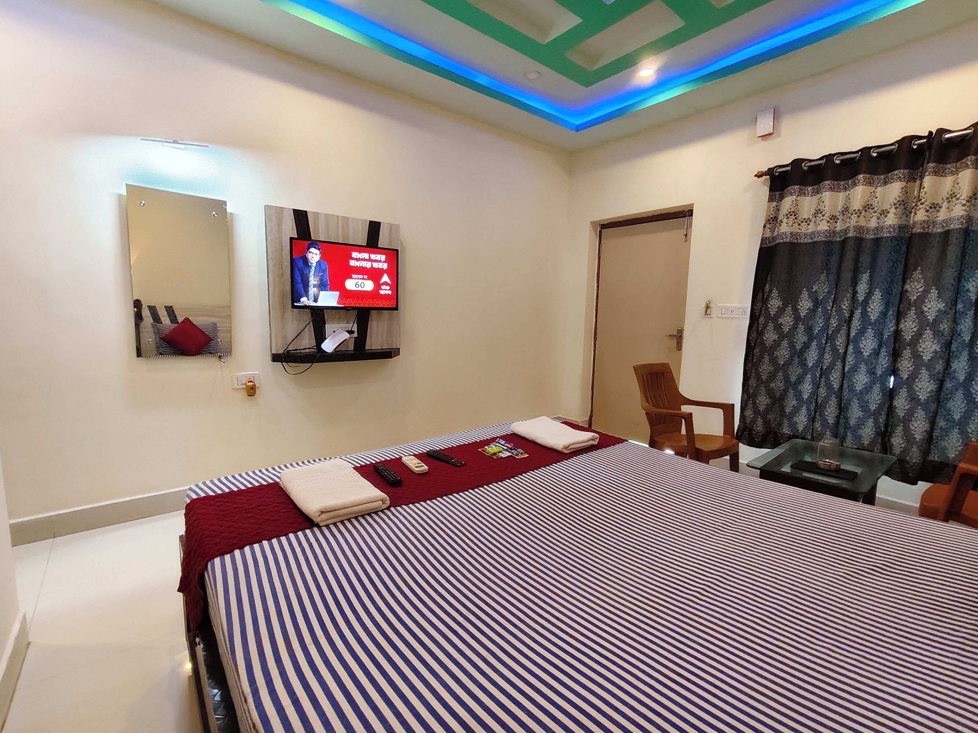 Una habitación de hotel con una cama, televisión montada en la pared, una silla y un ventanal con cortinas.