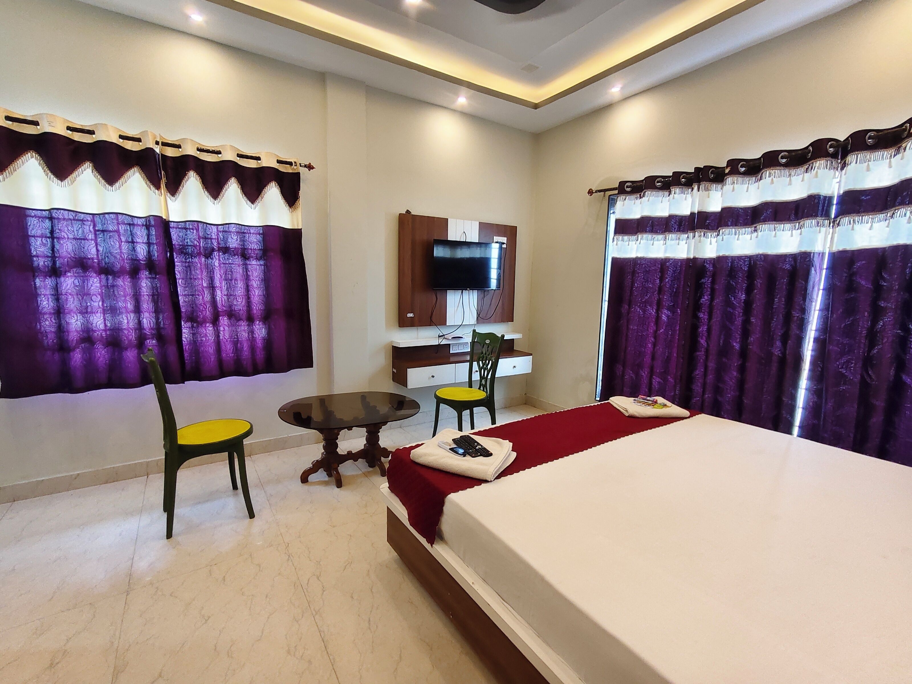 A hotel room with a bed, a desk with a chair, a television on the wall, and purple curtains.