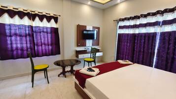 A hotel room with a bed, a desk with a chair, a television on the wall, and purple curtains.