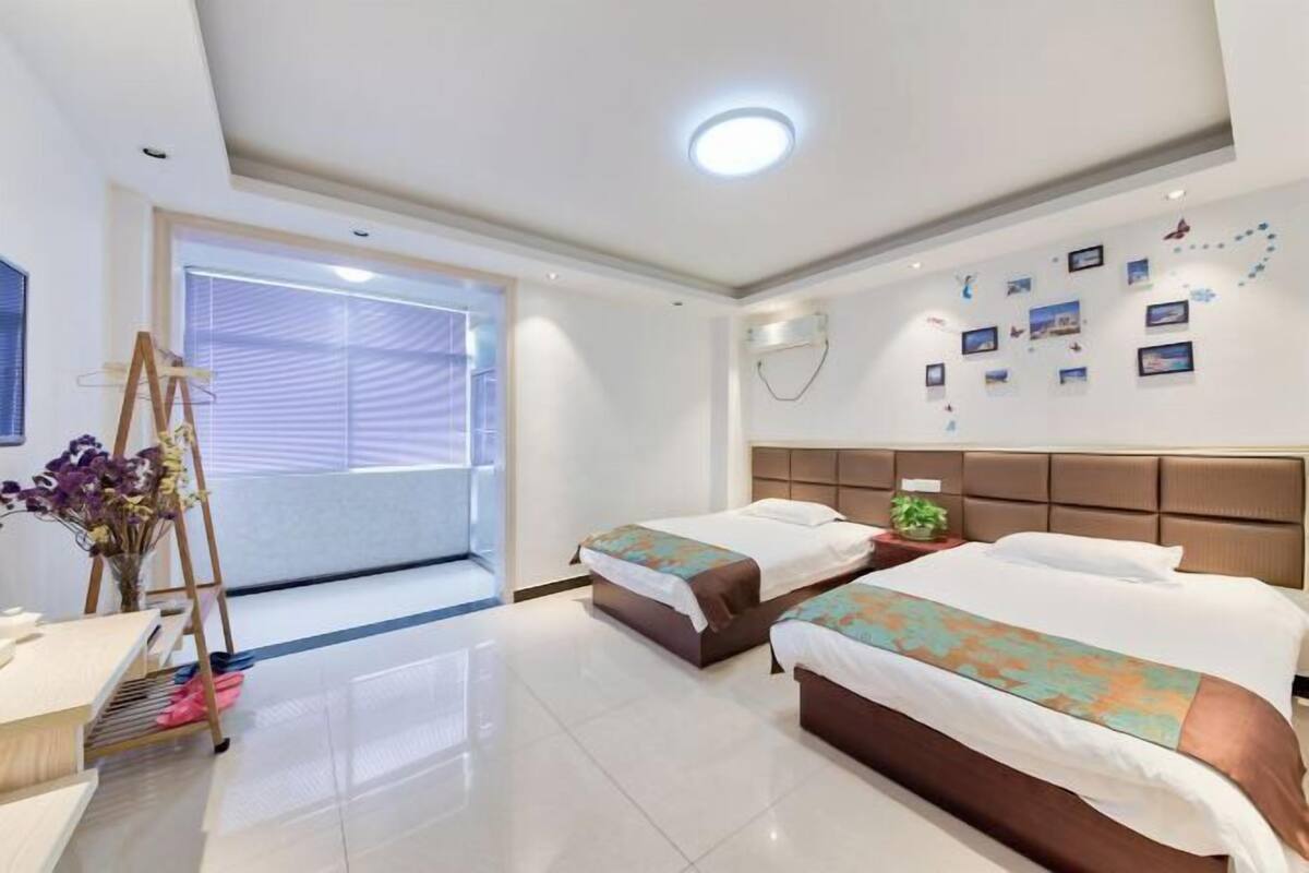 A hotel room with two beds, a desk, a vanity, and a large window with blinds.