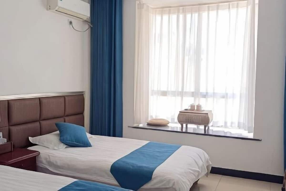 A hotel room with two beds, a window with sheer curtains, a bedside table with a lamp, and a wall-mounted air conditioner.