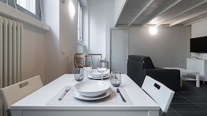 A dining area with a white table set for four, including plates, glasses, and cutlery. There is a black chair and a white cabinet with a television in the background. - Italianway Easy - Carcano 27 (Milano)