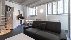 Ein modernes Wohnzimmer mit grauem Sofa, einer Wendeltreppe, einer Küchenzeile und großen Fenstern.