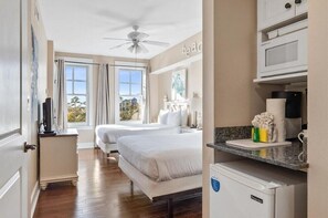 A hotel room with two beds, a kitchenette, a ceiling fan, and a view of the outdoors.