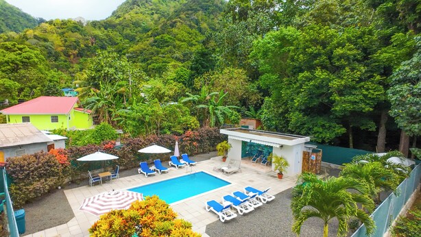 A pool area with lounge chairs and umbrellas, surrounded by lush greenery and a small building with a red roof. - Charming 1-bedroom apartment with WiFi, AC, swimming pool, in lovely Soufrière (St lucia)