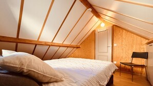 Ein Schlafzimmer mit schrägverdeckter Decke, Holzbalken, einem Bett mit weißer Bettwäsche, einem Sessel und einer Tür.