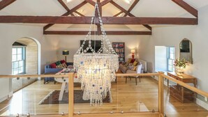 A spacious loft with exposed wooden beams, a large chandelier, a glass railing, and a cozy seating area with cushions and a bookshelf. - Stapleford Farm (Beaminster)