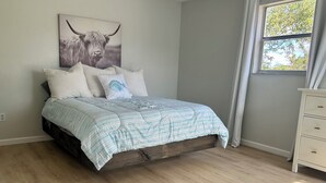 Une chambre à coucher avec un lit, une grande fenêtre avec des rideaux et une œuvre d’art représentant un taureau, accrochée au mur.