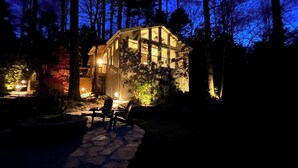 A cabin with a stone patio and chairs, surrounded by trees. - Cottage · Private Lake + Dock · Walk to Town (Cashiers)