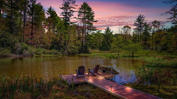 A wooden dock extending into a calm lake surrounded by dense forest, with chairs and a canoe on the dock. - Cottage · Private Lake + Dock · Walk to Town (Cashiers)