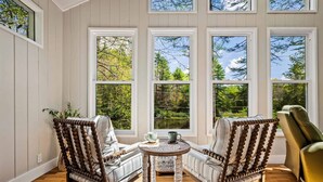 A cozy room with two upholstered chairs, a small round table, and large windows offering a view of trees and sky. - Cottage · Private Lake + Dock · Walk to Town (Cashiers)