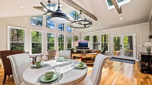 A spacious dining area with a round table set for four, surrounded by wooden flooring and large windows offering a view of the outdoors. - Cottage · Private Lake + Dock · Walk to Town (Cashiers)