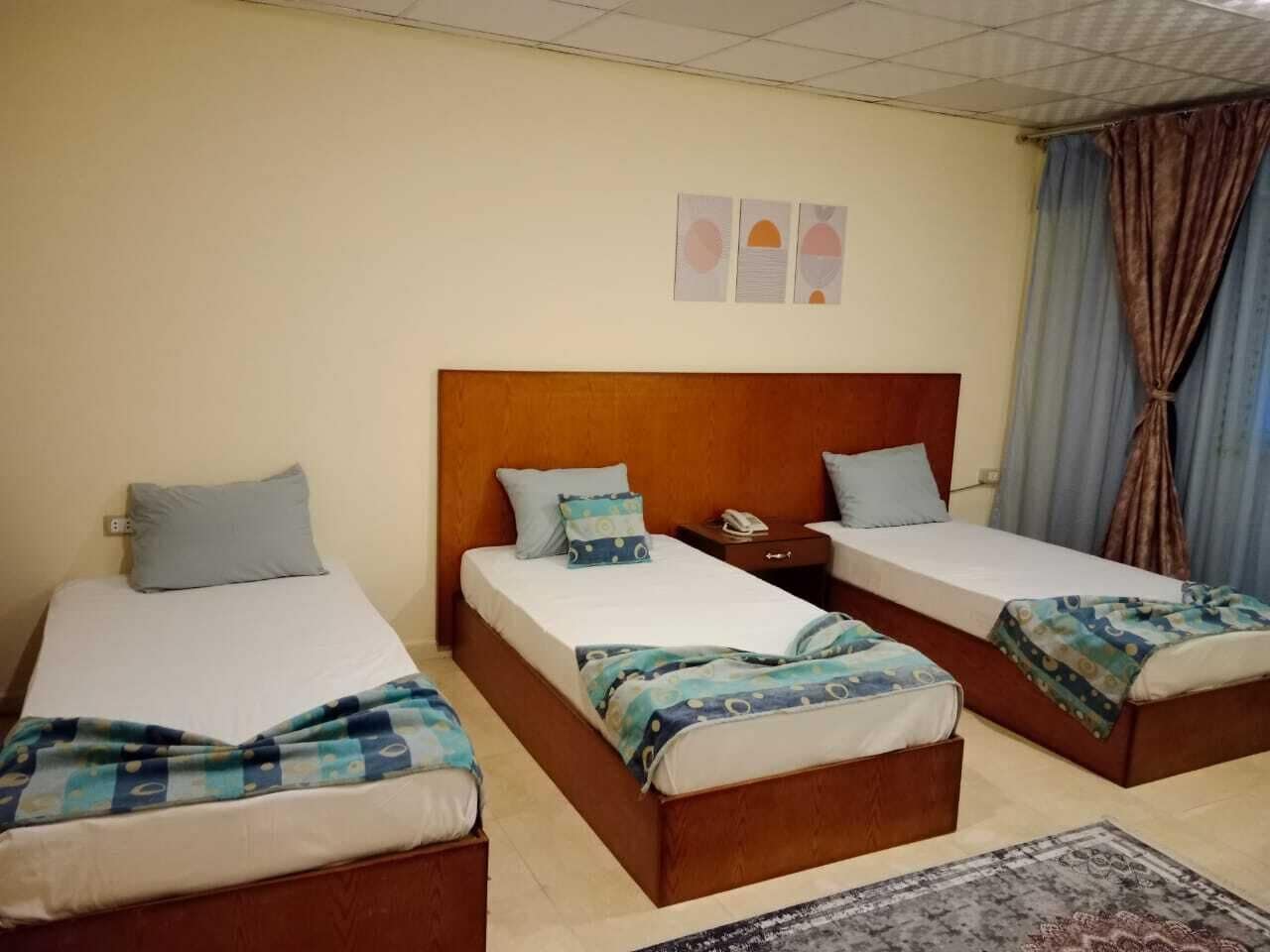 A hotel room with three single beds, each with a wooden headboard, a bedside table, and a telephone. The room has a tiled floor and a patterned rug.
