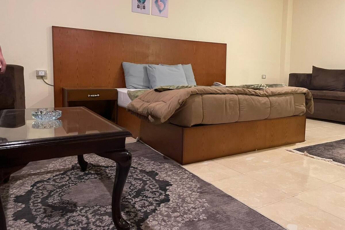 A bedroom with a wooden headboard, a bed with a beige and brown blanket, a glass coffee table, and a dark patterned rug.