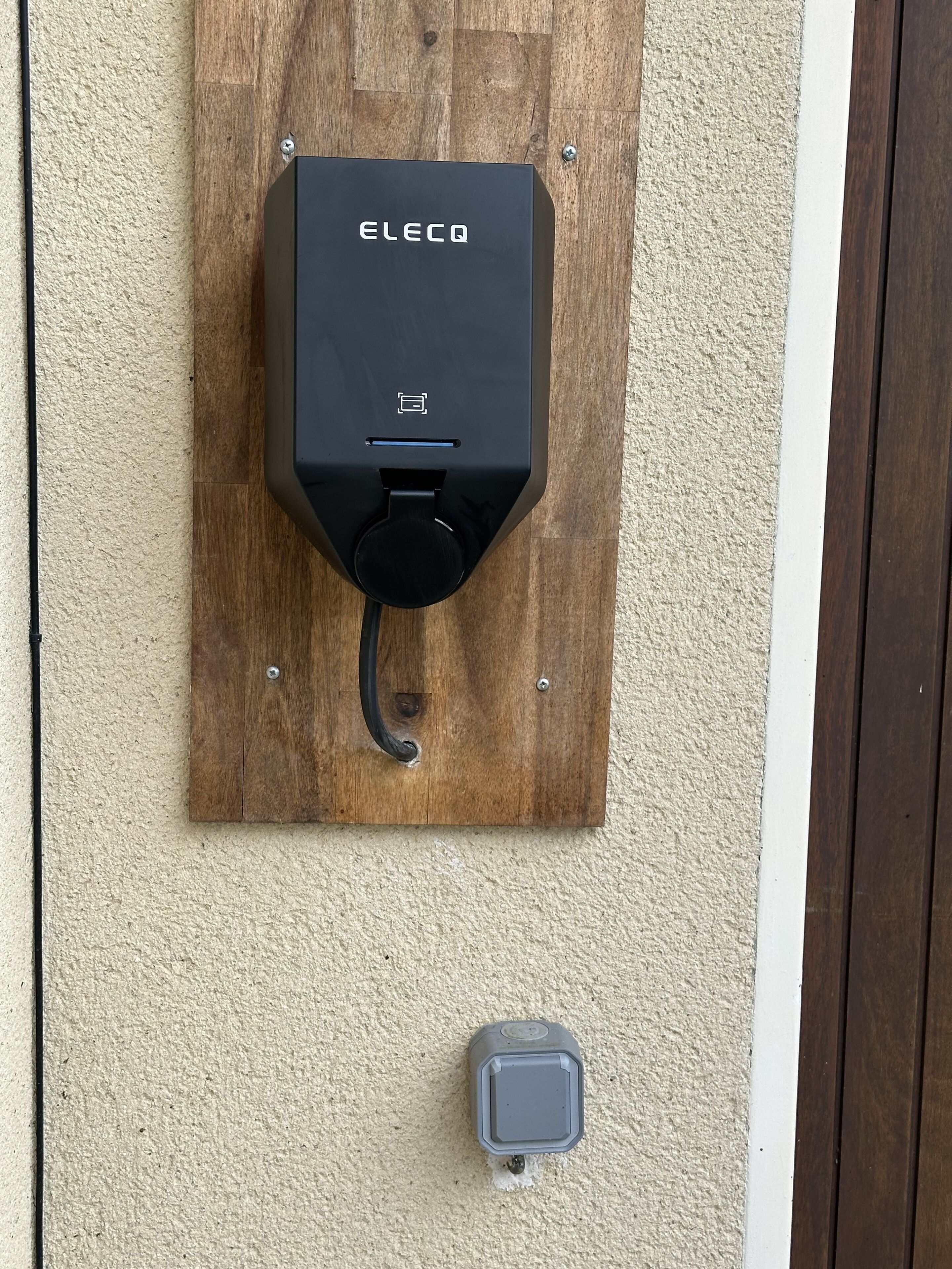 A black ELECQ charging station mounted on a wooden board attached to a textured wall.
