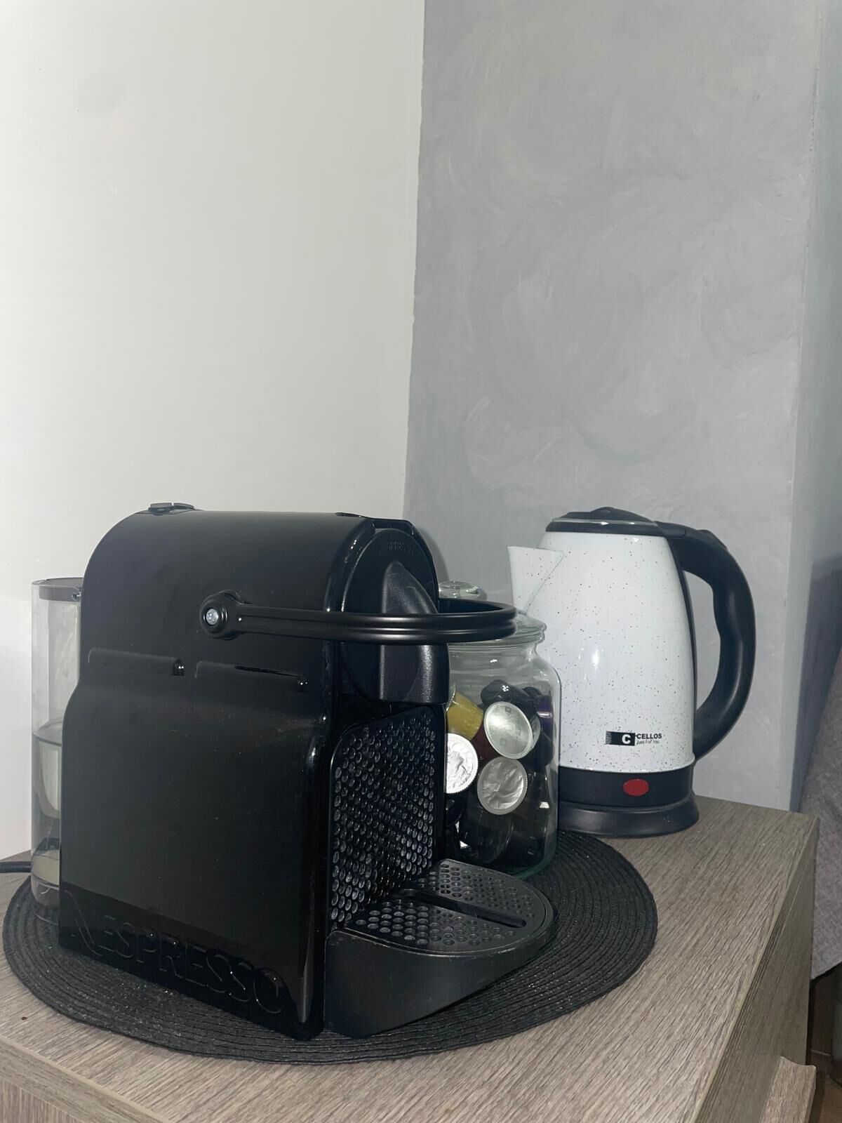 Een zwarte espresso machine op een ronde mat, een witte elektrische waterkoker, en een glazen pot met muntstukken op een houten tafel.