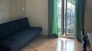 Une pièce avec un canapé bleu foncé, un sol carrelé et une porte-fenêtre à glissière avec des rideaux verts.