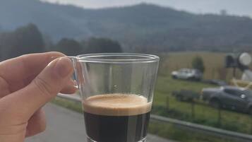 A hand holding a glass of dark liquid, likely coffee, with a scenic view of a road and mountains in the background.